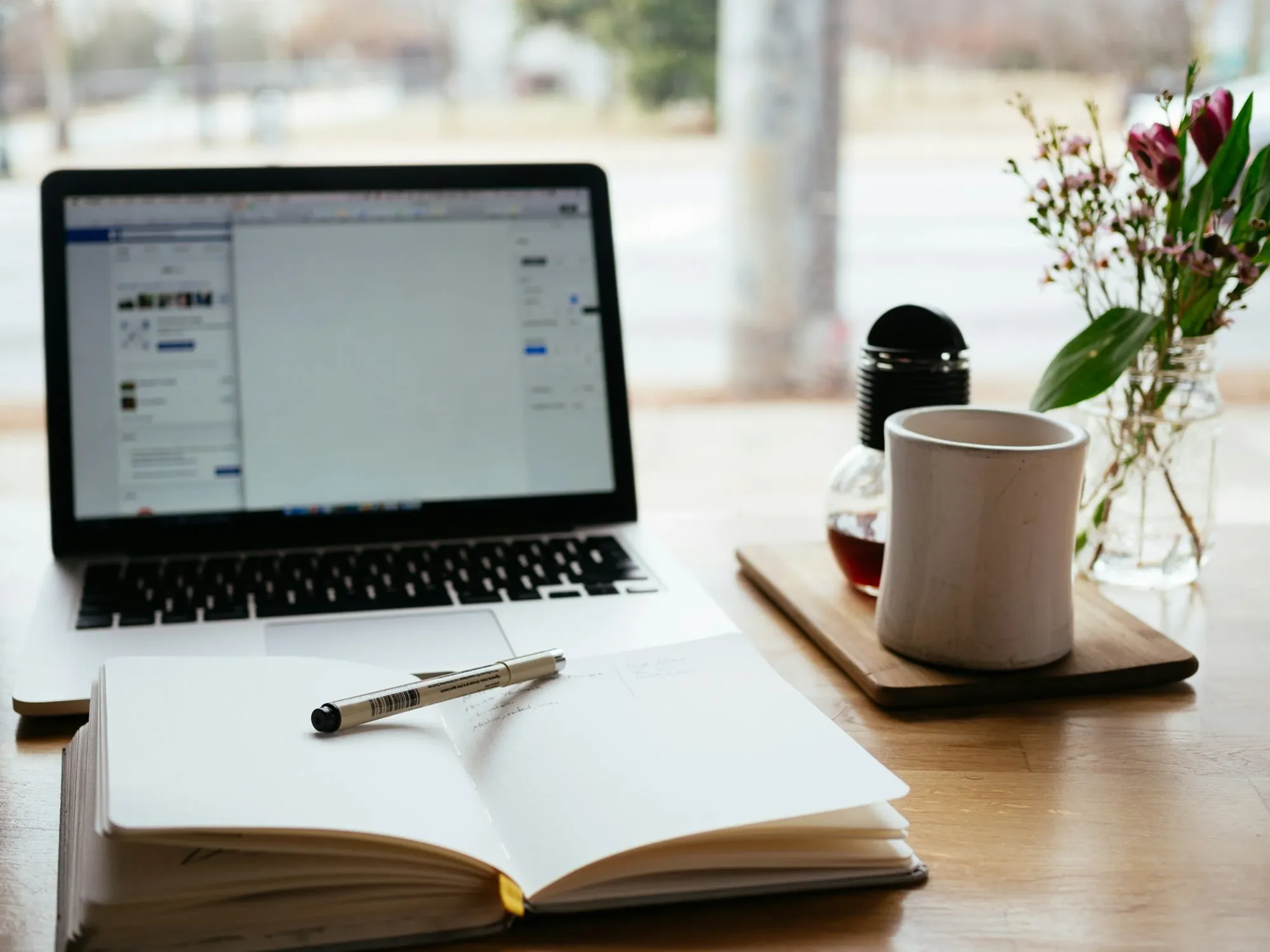 Aufgeschlagenes Notizbuch mit Stift vor einem Laptop, daneben ein Becher und Blumen in einer Glasvase